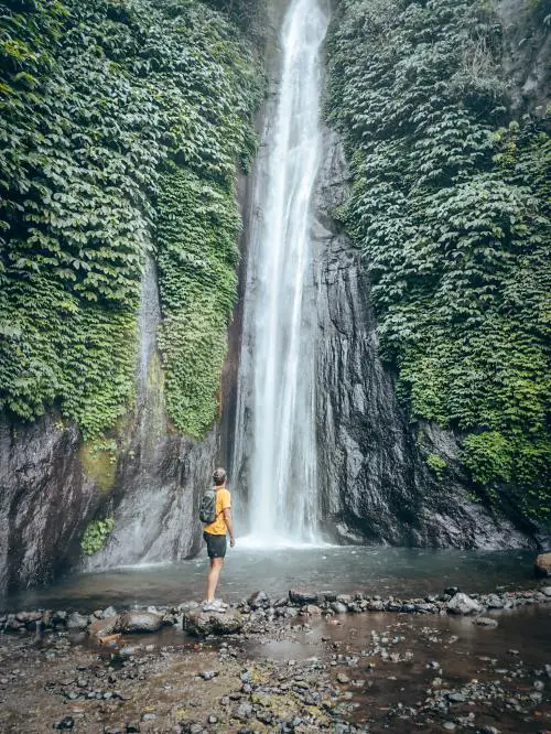 A man wearing a yellow t-shirt faces the Red Coral Waterfall (Munduk Waterfall) in Bali, Indonesia. This is one of the best waterfalls to visit in Bali!