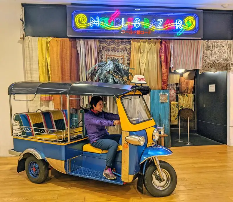 Jackie Szeto from Life Of Doing rides a yellow and blue tuk tuk in front of the Cup Noodles Museum's Noodles Bazaar. It's a spot where people can try mini bowls of noodles from around the world.
