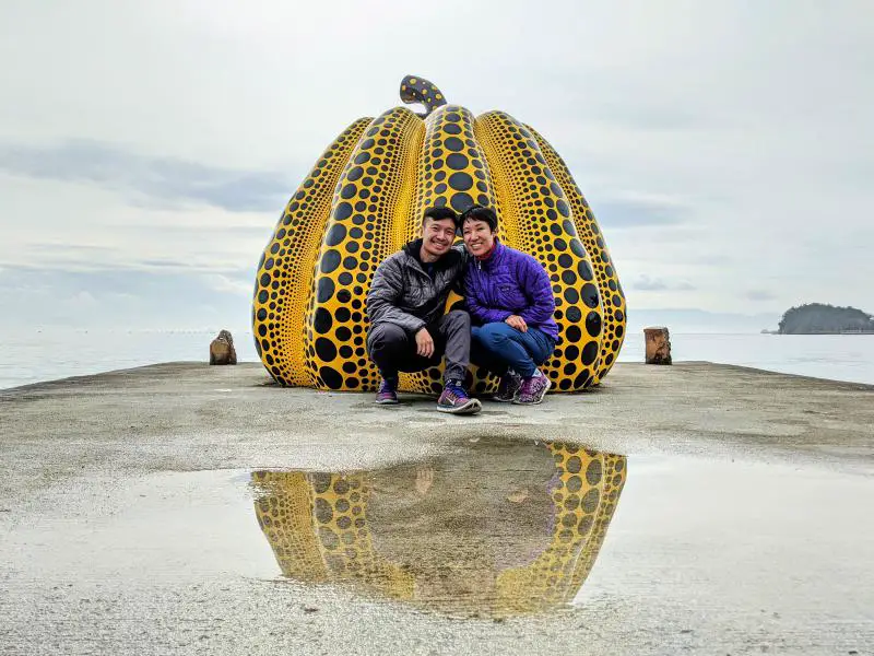 Justin Huynh and Jackie Szeto from Life Of Doing poses in front of Yellow Pumpkin sculpture created by Yayoi Kusama. The sculpture is a top attraction to visit on Naoshima, Japan.