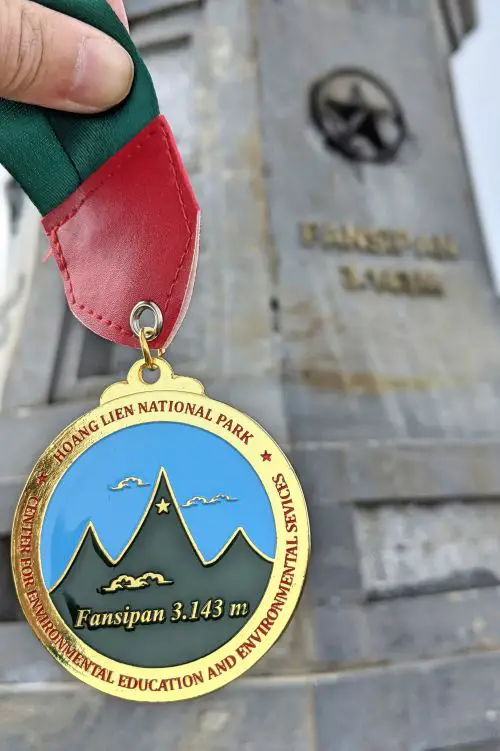 Mount Fansipan hike finisher's medal in front of the Fansipan summit sign in Sapa, Vietnam