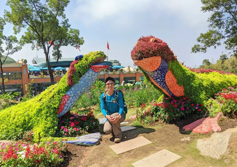 Jackie Szeto, Life Of Doing, pose underneath two lizard topiaries made out of green and red plants at Sun World Ba Den in Vietnam