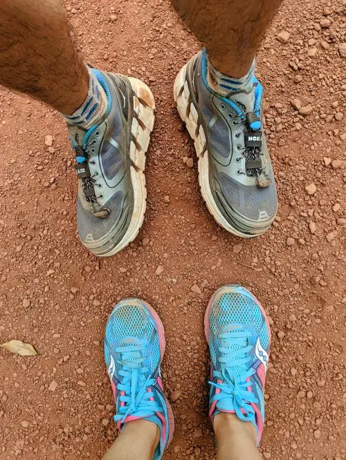 An aerial view of a pair of black and blue Hoka running shoes and a pair of pink and baby blue Saucony running shoes on the red clay dirt trail in Ma Da Forest, Vietnam
