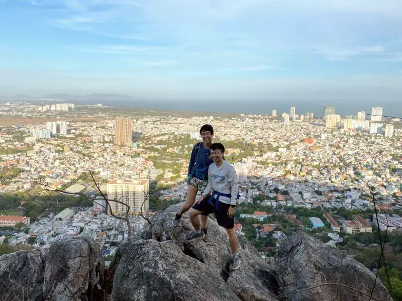 Justin Huynh and Jackie Szeto, Life Of Doing, stand on a rock with the Vung Tau City and buildings in the background on Nui Lon Big Mountain, Vietnam
