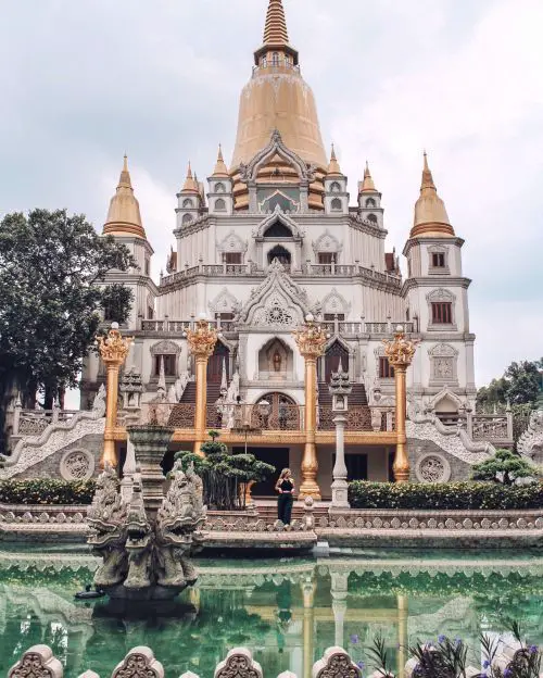 A woman in a black dress stands in front of a white and gold Buddhist Pagoda, the Buu Long Pagoda, in Ho Chi Minh City, Vietnam