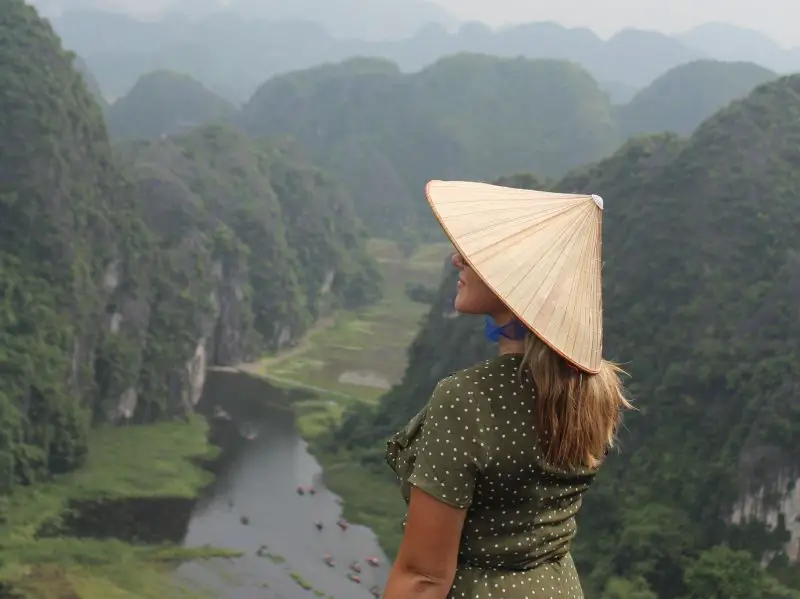 When packing for Vietnam, add comfortable clothes to your luggage. A woman wears a green and white polka dot dress and a conical hat and overlooks Ninh Binh landscapes.