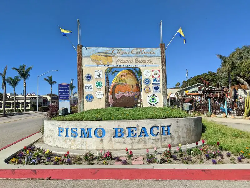 Pismo Beach sign with Pismo Beach service club logos and a painting of a palm tree and ocean.