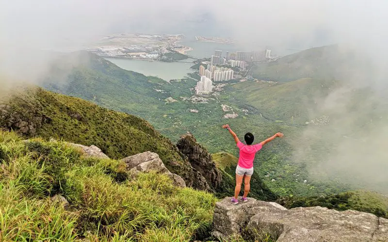 Jackie Szeto, Life Of Doing, in a pink shirt overlooks an apartment area on top of the Lantau Peak hike in Hong Kong