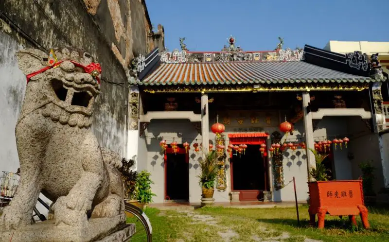 A stone lion statue with a red ribbon is in front of a temple in Penang, Malaysia