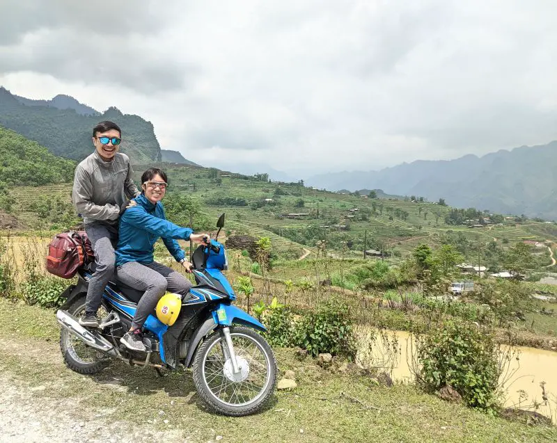 Jackie Szeto and Justin Huynh, Life Of Doing, sit on a top of a motorbike with green rice fields and mountains in Ha Giang, Vietnam