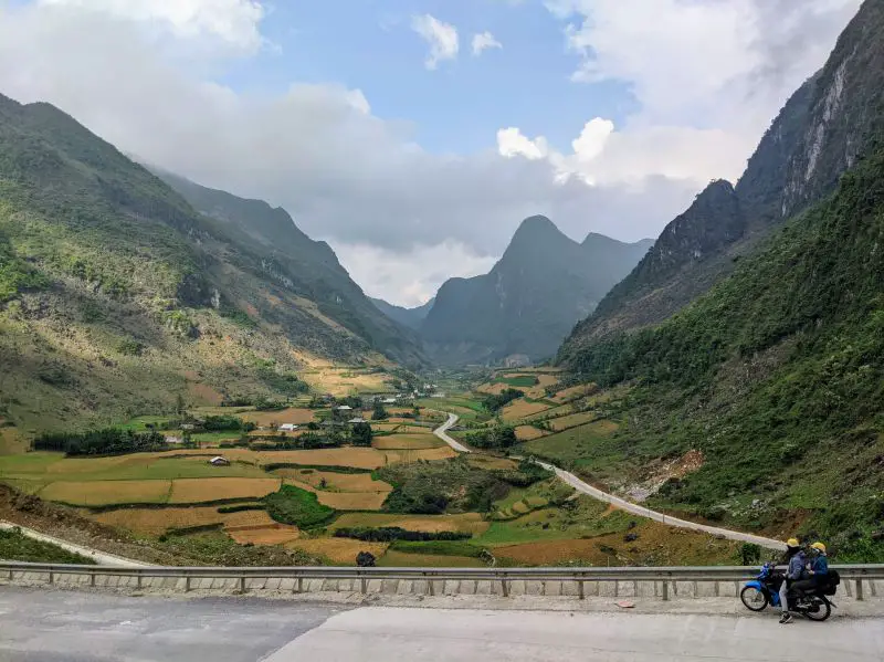 Two people on a motorbike overlook a valley with tall mountains and rice fields in Cao Bang, Vietnam