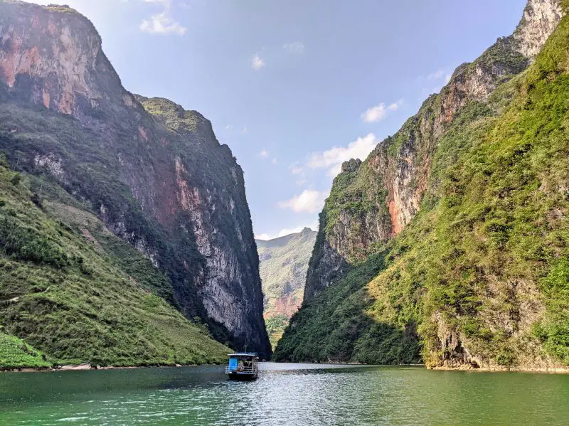 A boat rides along the turquoise Nho Que River and passing by two tall mountains.