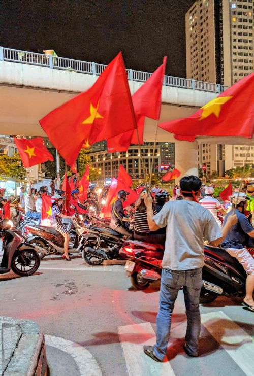 A swarm of motorbikes on the road and people waving the Vietnam flag which is red and has a yellow star in the middle