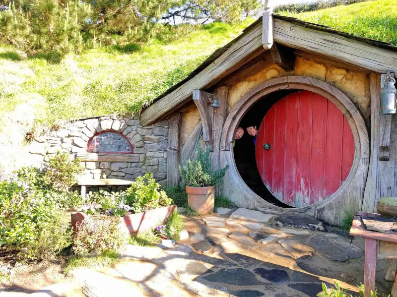Justin Huynh and Jackie Szeto, Life Of Doing, peek their heads behind the red Hobbit hole door at Hobbiton Movie Set.