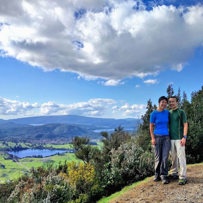 Jackie Szeto and Justin Huynh, Life Of Doing, at the summit of Rainbow Mountain in Rotorua, New Zealand and overlooking a small lake and mountains.