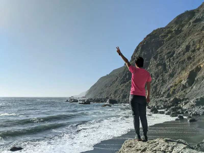 Jackie Szeto, Life Of Doing, holds a peace sign and overlooks the ocean at Ragged Point Trail, Ragged Point, California