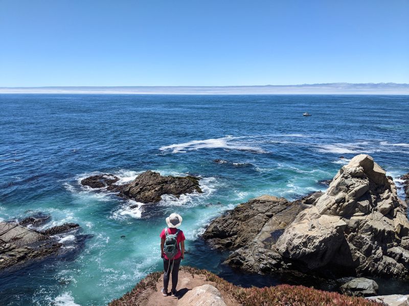 Jackie Szeto, Life Of Doing, stands on a dirt trail and overlooks the blue and teal Pacific Ocean