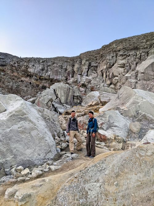 Justin Huynh and Jackie Szeto, Life Of Doing, stand on the Ijen hiking trail with tall grey rocks in the back
