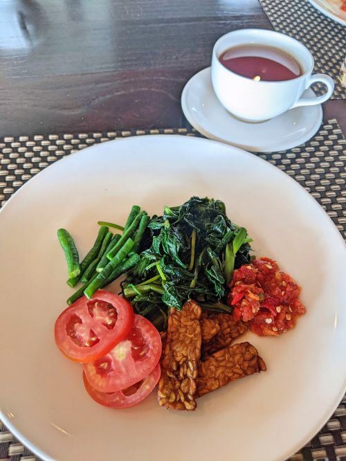 A plate of sliced tomatoes, green beans, green leafy vegetables, fried tempeh, and sambal at Ketapang Indah Hotel's breakfast