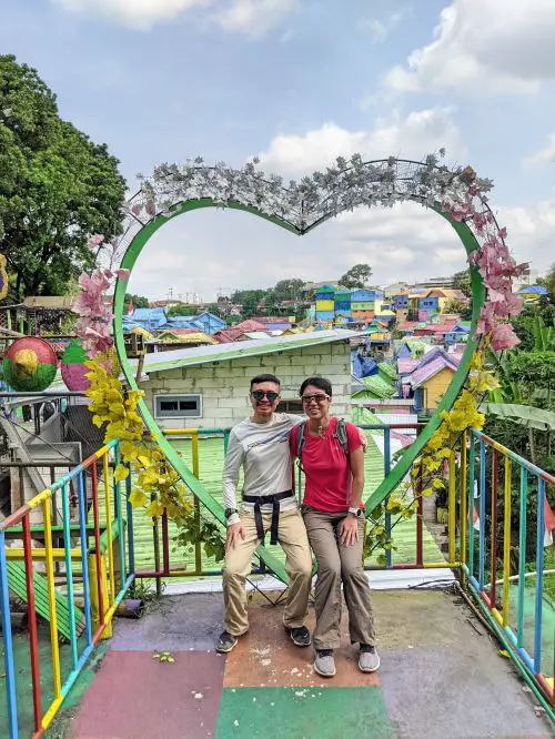 Justin Huynh and Jackie Szeto, Life Of Doing, sit with in heart shape prop with the colorful village in the back at Malang, Indonesia
