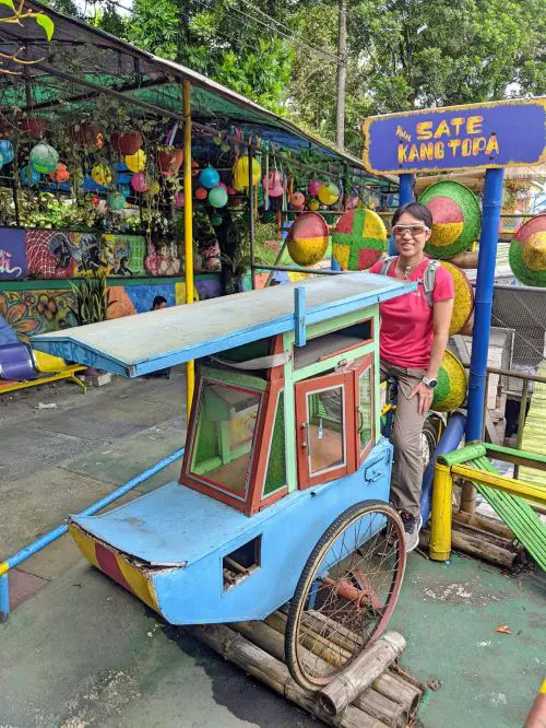 Jackie Szeto, Life Of Doing, interacts with a 3-D prop and sits on a bicycle seat pretending to sell satay at Wisata Kampung Tridi, Malang, Indonesia