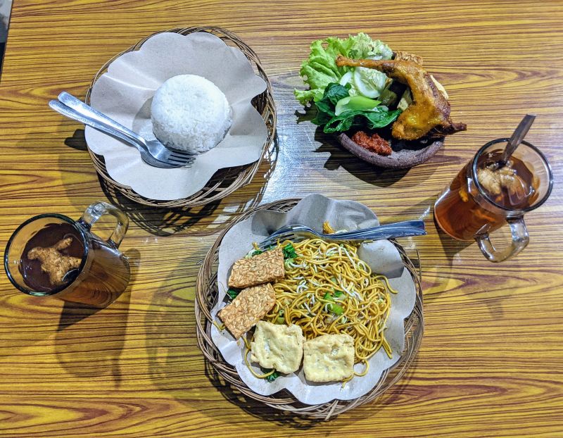 A plate of fried chicken leg with greens and rice and a plate of fried noodles with tempeh and tofu and two glasses of tea