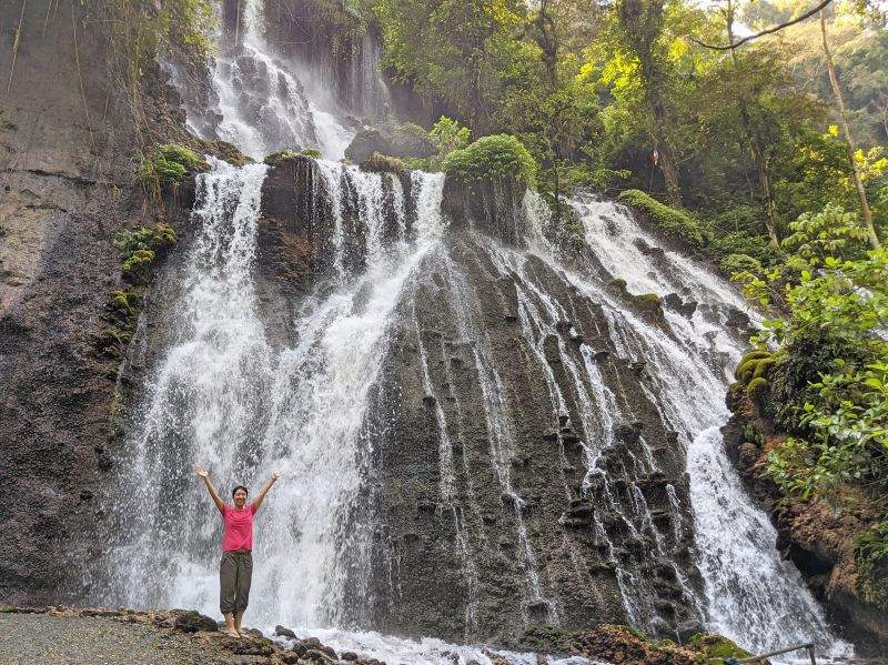 Jackie Szeto, Life Of Doing, has arms up and stands next to Goa Tetes Waterfall, Indonesia