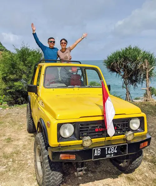 Justin Huynh and Jackie Szeto, Life Of Doing, stand in the back of a yellow Jeep and hold their hands up at Timang Beach, Indonesia
