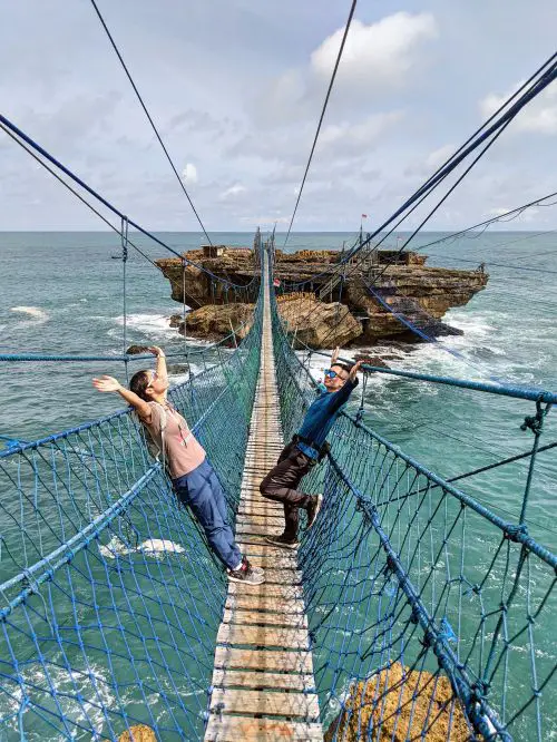 Jackie Szeto and Justin Huynh, Life Of Doing, are on a suspension bridge over the ocean and holding their hands up at Timang Beach area, Indonesia