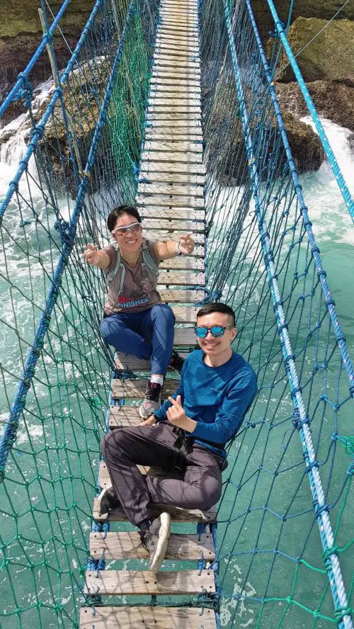 Jackie Szeto and Justin Huynh, Life Of Doing, are on Timang Beach suspension bridge over the ocean and holding fingers in a heart shape