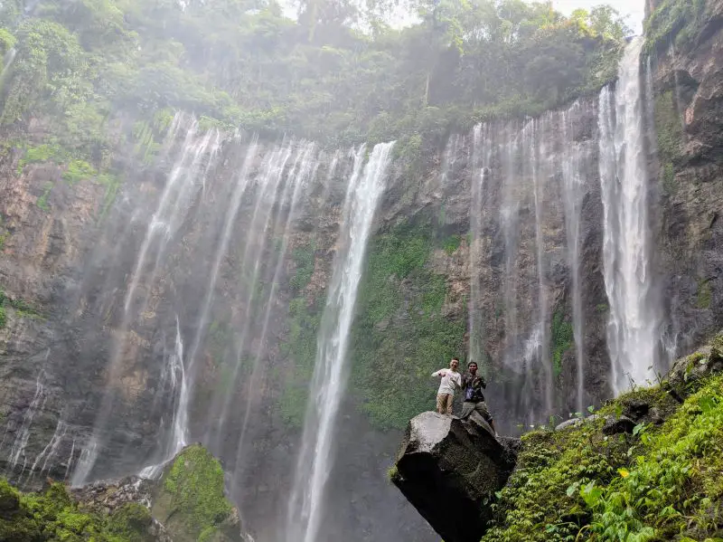 Justin Huynh, Life Of Doing, and Arief, tour guide, stand on a rock with Tumpak Sewu Waterfall cascades in the background