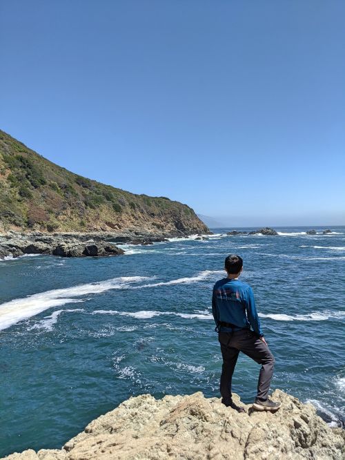 Justin Huynh, Life Of Doing, stands on a rock and looking at the blue ocean waters from Partington Cove, Big Sur, California