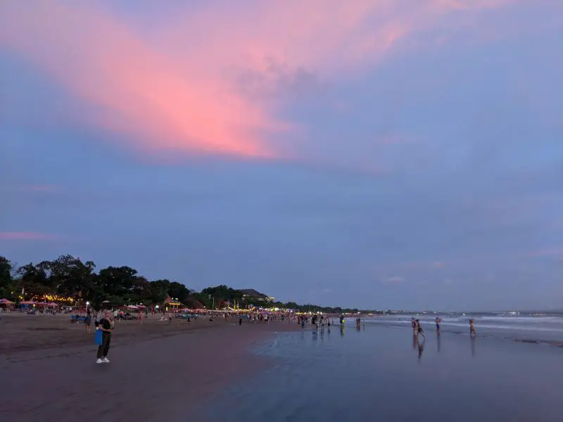 People walk on Seminyak's black sand beach with the pink clouds at sunset