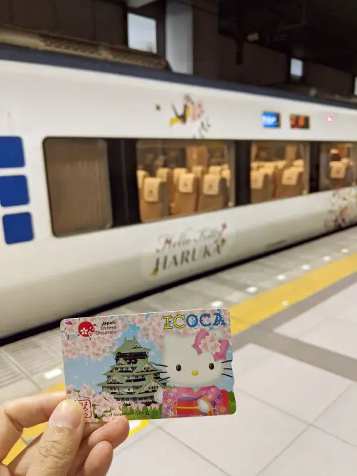 A hand holding up a Hello Kitty ICOCA card in front of the Hello Kitty Haruka train at Kansai Airport Station