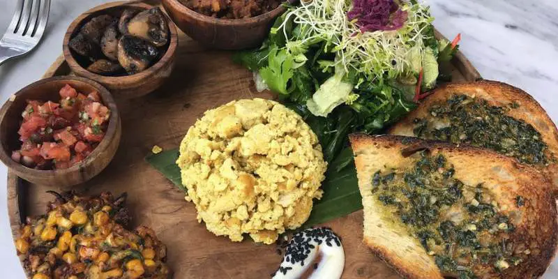 A wooden platter with toasted bread, salad, mushrooms, salsa, corn, vegan egg, and other vegan items at Pels Supershop in Canggu