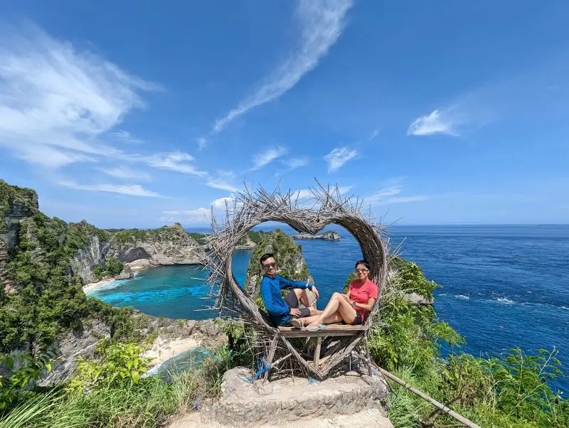 Justin Huynh and Jackie Szeto, Life Of Doing, sit in a heart shaped wooden seat at one of the photo spots at Thousand Islands Viewpoint in Nusa Penida
