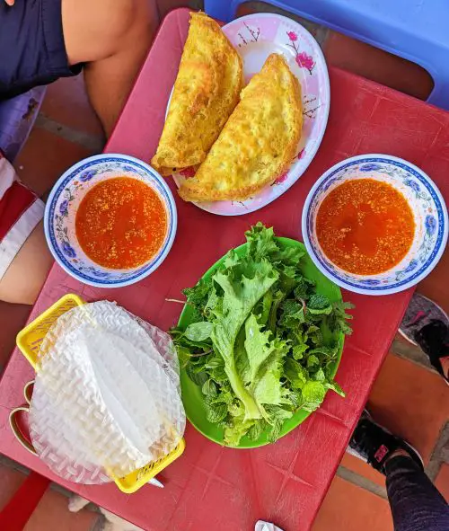 A plate of Vietnamese sizzling pancake, two bowls of red fish sauce, a plate of lettuce and herbs, and rice wrappers at Banh Xeo Co Bay, Phu Quy