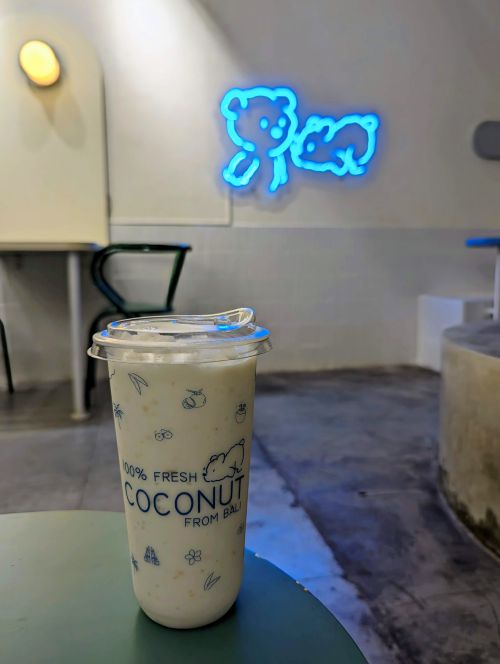 A large cup of original coconut shake with the neon light bear logo on the wall at Kookos Cafe, Seminyak