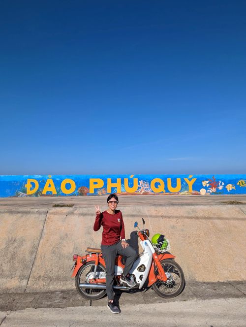 Jackie Szeto, Life Of Doing, sit on an orange and white motorbike with Dao Phu Quy street art in the background