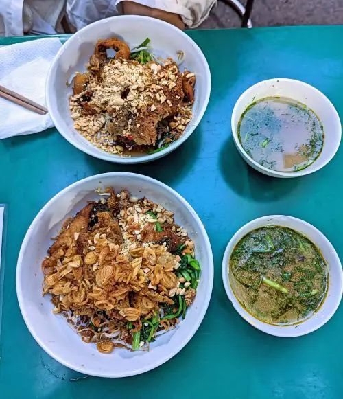 Two bowls of fried fish noodles topped with fried fish, green vegetables, fried onions, and peanuts, and two bowls of soup with dill on the side at Bun Ca Sam Cay Si in Hanoi