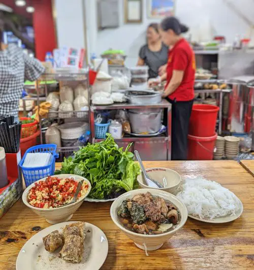 A table with a plate of fried crab pork spring roll, a bowl of pork meatballs, a plate of noodles, a plate of fresh vegetables and herbs, and a bowl of diced chili at Bun Cha Dac Kim in Hanoi