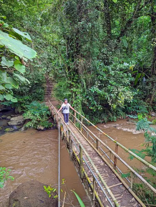 An older woman holding the bridge railings to cross over a river at Leke Leke Waterfall