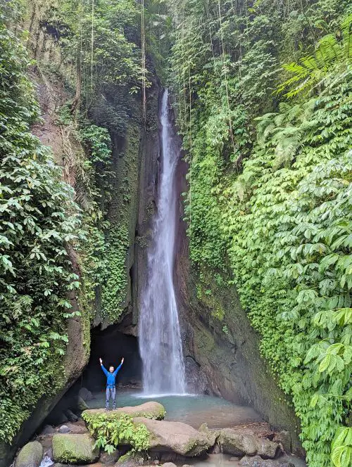 Justin Huynh, Life Of Doing, stands on a rock with hands in the air and has the Leke Leke Waterfall behind him