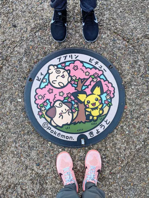 Two people's feet stand in next to the Pokemon manhole cover with a design of three Pokemon characters and cherry blossom trees in Kyoto, Japan