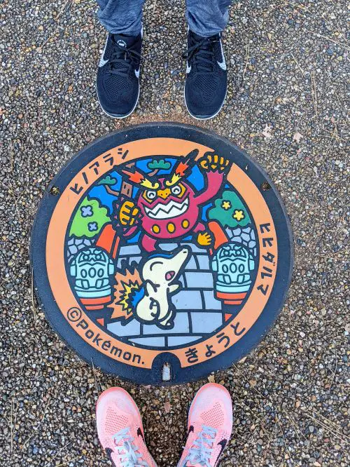 Two people's feet stand next to the Pokemon manhole cover with a design of two Pokemon characters and a shrine