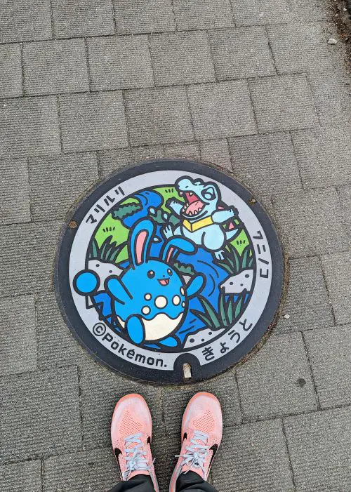 A person's feet stands next to the Pokemon themed manhole cover and the design has two Pokemon characters and a river