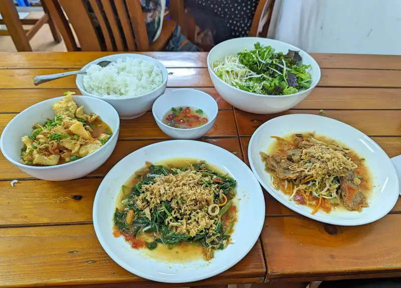 Six bowls of vegetarian food such as faux meat, morning glory vegetables, tofu, rice, and raw vegetables at Sen Trang An Vietnamese Restaurant in Hanoi