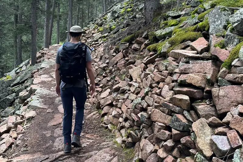 A male hiker wearing a backpack and hat and walking on a rocky hiking trail next to a wall of rocks