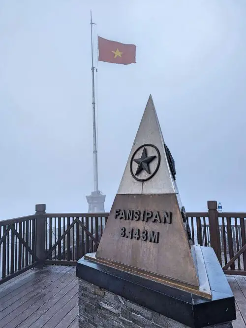 A triangular landmark that says Fansipan 3,143 meters and the Vietnam flag waving on a flagpole at Fansipan peak in Sapa, Vietnam