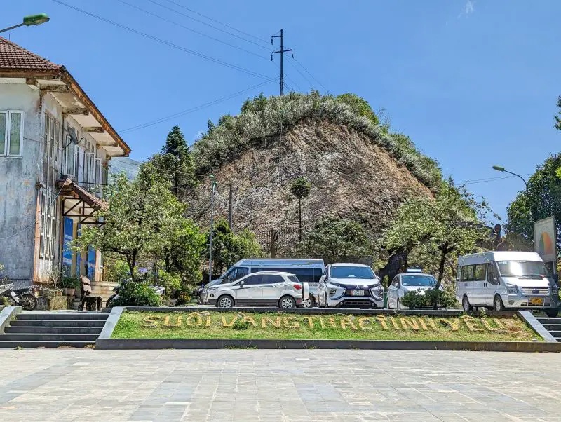 A parking lot with cars and vans and a sign of Suối Vàng Thác Tình Yêu, which translates to Love Waterfall Golden Stream in Vietnamese