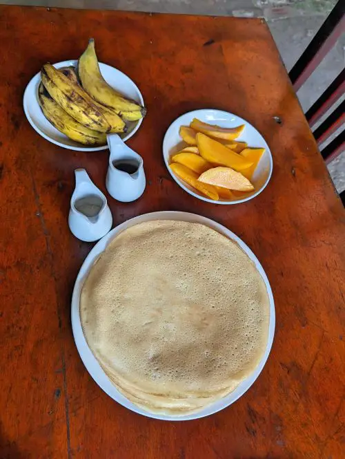 A plate of crepes with bowls of fresh cut mangoes and whole bananas on a wooden table at May's Homestay in Sapa
